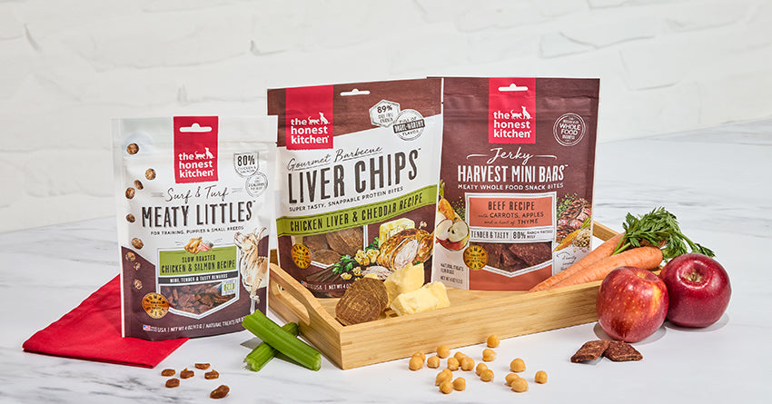 The Honest Kitchen's line of meaty treats for dogs on a marble counter surrounded by ingredients.