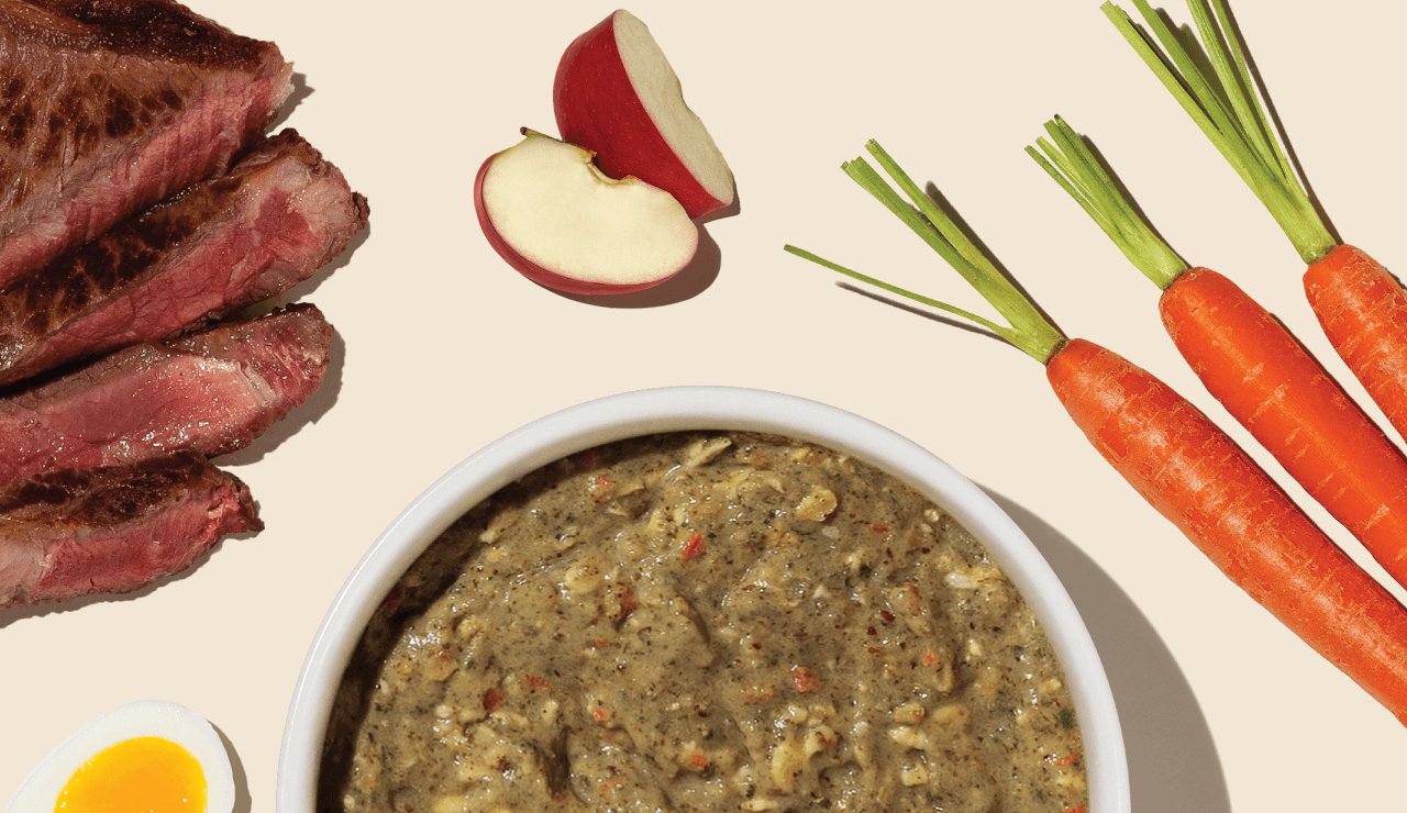 A top-down view of a balanced meal featuring steak slices, apple slices, carrots, a bowl of stew, and an egg half on a light surface. Shop healthy, high quality, human grade dehydrated dog food from The Honest Kitchen.