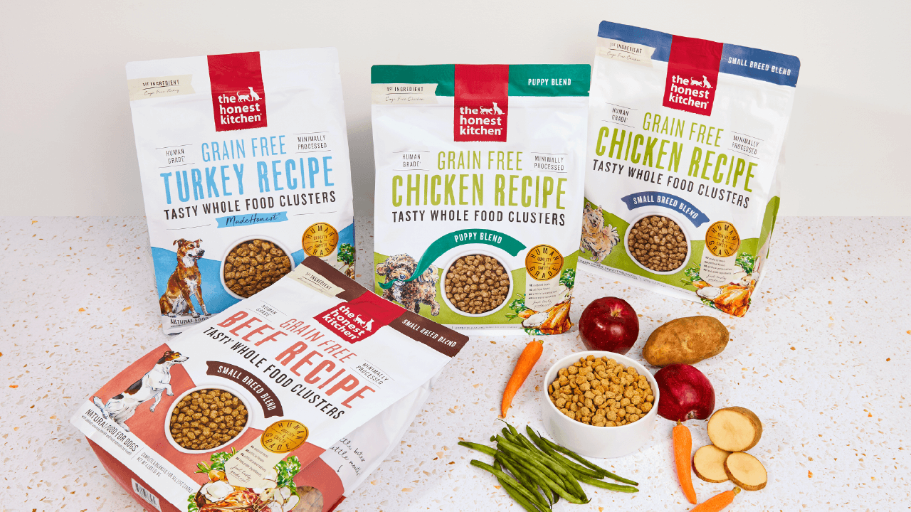Four bags of The Honest Kitchen Grain Free dog food, including Turkey, Chicken, and Beef recipes, are showcased with fresh ingredients like carrots, green beans, apples, and potatoes.