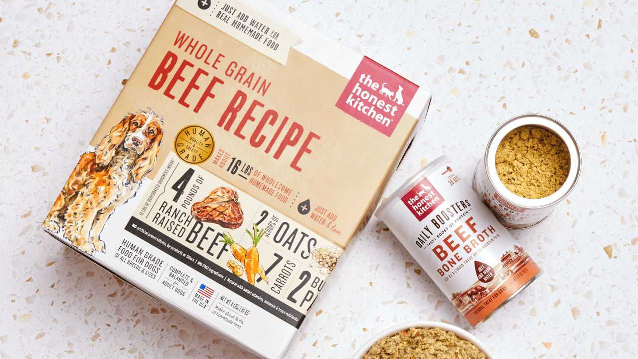 The Honest Kitchen Whole Grain Beef Recipe dog food and a bone broth booster are shown on a speckled surface. Shop beef dog food, treats, and toppers from The Honest Kitchen. 