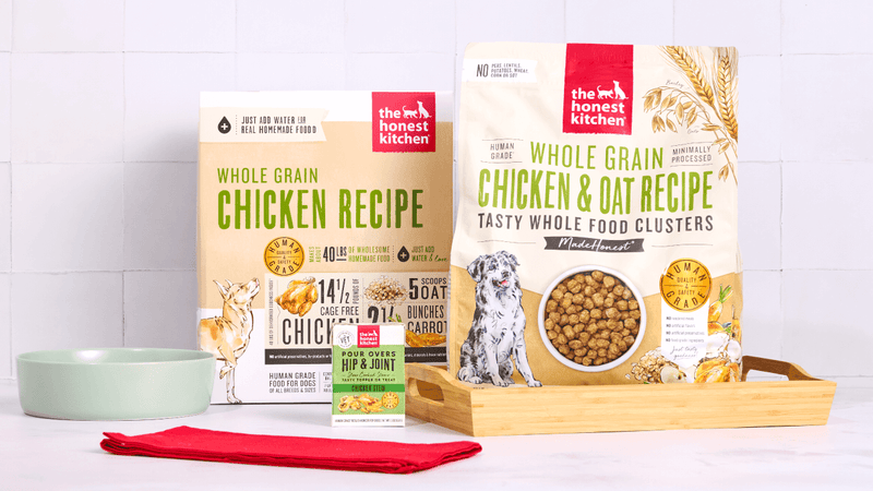 Two bags of The Honest Kitchen Whole Grain Chicken dog food are displayed with a bowl, red cloth, and a supplement box. Shop dog best sellers from The Honest Kitchen.