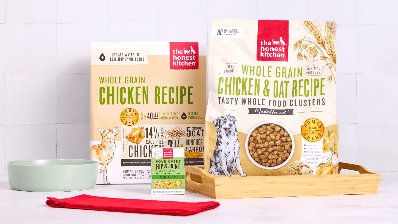 Two bags of The Honest Kitchen Whole Grain Chicken dog food are displayed with a bowl, red cloth, and a supplement box. Shop dog best sellers from The Honest Kitchen.