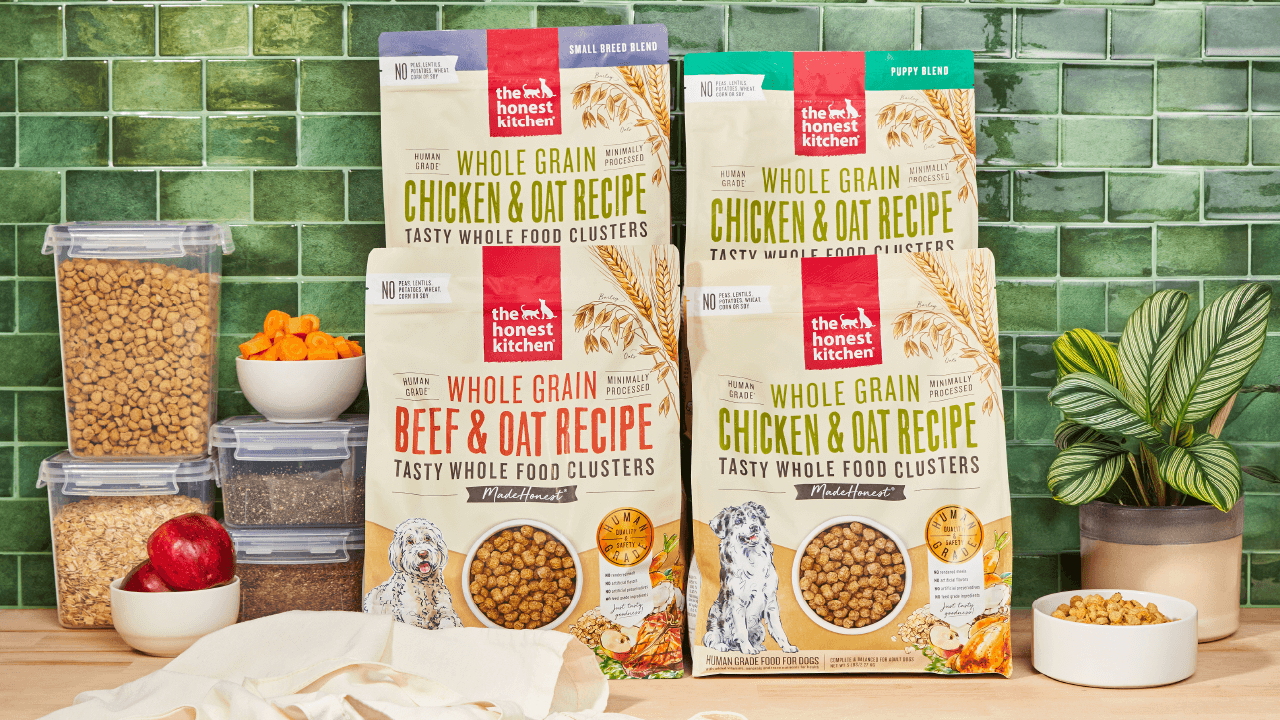 A product shot featuring multiple bags of The Honest Kitchen Whole Grain dog food, including Chicken & Oat and Beef & Oat recipes, displayed with fresh ingredients and storage containers.