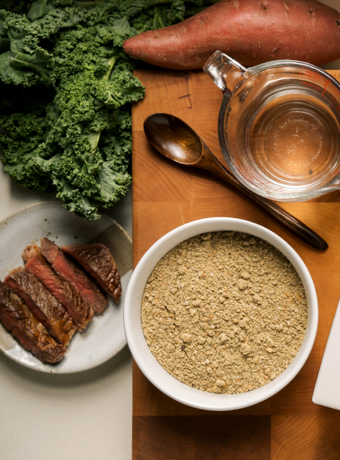 Bowl of dehydrated food with kale, sweet potato, and sliced beef on a wooden board. About the honest kitchen.