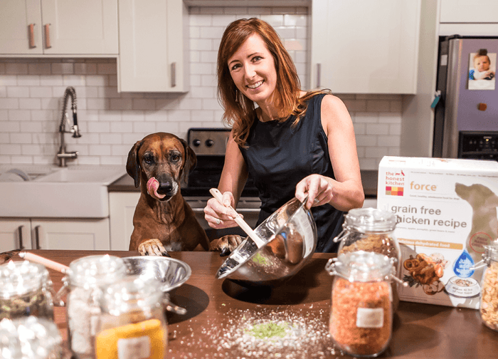Image of The Honest Kitchen's Founder Lucy Postins in a kitchen with ingredients, her dog and an old box of The Honest Kitchen. About The Honest Kitchen.